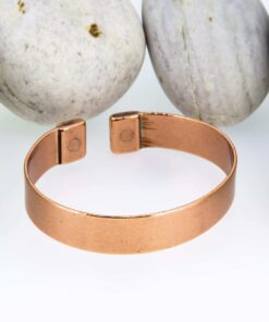 Plain polished magnetic copper therapy bangle with a smooth wide band and rectangular magnetic ends, displayed against a backdrop of smooth white pebbles.