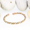 Slim twisted magnetic copper therapy bangle combining copper, brass, and silver-tone strands, finished with round magnetic ball ends, displayed in front of smooth white pebbles.