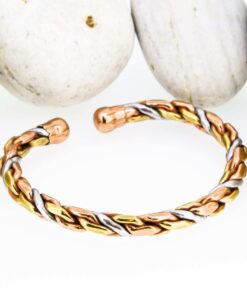 Braided magnetic copper therapy bangle with intertwined copper, brass, and silver-tone strands, finished with round magnetic ball ends, displayed in front of smooth white pebbles.