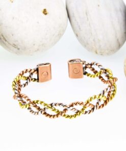Open magnetic copper therapy bangle with twisted copper, brass, and silver-tone wires in an interlinked design, featuring rectangular magnetic ends, displayed against smooth white pebbles.