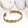 Braided magnetic copper therapy bangle combining copper, brass, and silver-tone strands, with rectangular magnetic ends, displayed against a backdrop of smooth white pebbles.