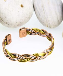 Braided magnetic copper therapy bangle combining copper, brass, and silver-tone strands, with rectangular magnetic ends, displayed against a backdrop of smooth white pebbles.