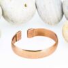 Textured magnetic copper therapy bangle with a fine lined pattern on a wide band, finished with rectangular magnetic ends, displayed against smooth white pebbles.