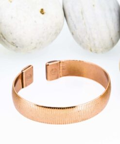 Textured magnetic copper therapy bangle with a fine lined pattern on a wide band, finished with rectangular magnetic ends, displayed against smooth white pebbles.