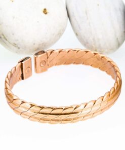 Braided-pattern magnetic copper therapy bangle with a textured, plaited design on a wide band, finished with rectangular magnetic ends, displayed against smooth white pebbles.