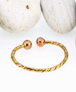 Slim twisted magnetic copper therapy bangle with intertwined copper and brass strands, finished with round magnetic ball ends, displayed against smooth white pebbles.