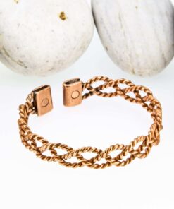 Braided-pattern magnetic copper therapy bangle with a textured, plaited design on a wide band, finished with rectangular magnetic ends, displayed against smooth white pebbles.