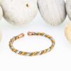 Slim twisted magnetic copper therapy bangle combining copper, brass, and silver-tone strands, finished with round magnetic ball ends, displayed in front of smooth white pebbles.