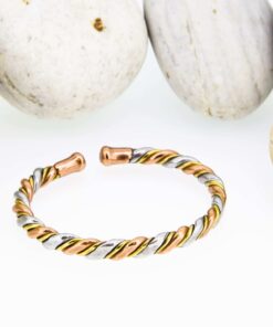 Slim twisted magnetic copper therapy bangle combining copper, brass, and silver-tone strands, finished with round magnetic ball ends, displayed in front of smooth white pebbles.