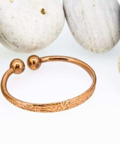 Copper magnetic therapy bangle with engraved floral detailing, featuring two polished magnetic ball ends, displayed in front of smooth white pebbles.