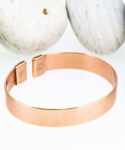 Plain polished magnetic copper therapy bangle with a smooth wide band and rectangular magnetic ends, displayed against a backdrop of smooth white pebbles.