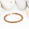 Slim twisted magnetic copper therapy bangle with a rope-style design and round magnetic ball ends, displayed in front of smooth white pebbles.
