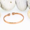 Slim polished magnetic copper therapy bangle with a smooth plain band and rectangular magnetic ends, displayed against a backdrop of smooth white pebbles.
