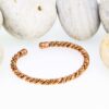 Slim twisted magnetic copper therapy bangle with a rope-style design and round magnetic ball ends, displayed in front of smooth white pebbles.