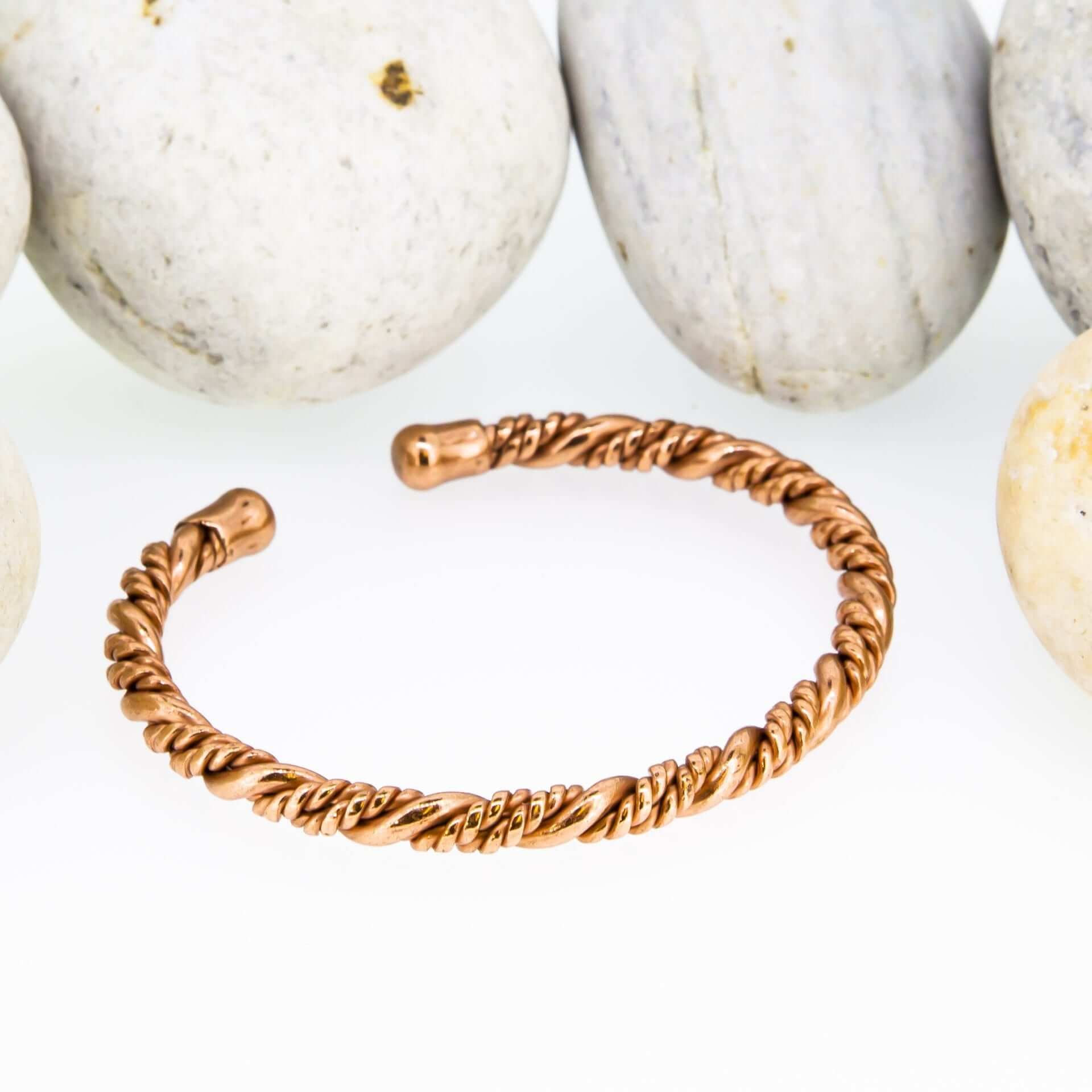 Slim twisted magnetic copper therapy bangle with a rope-style design and round magnetic ball ends, displayed in front of smooth white pebbles.