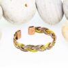 Braided magnetic copper therapy bangle combining copper, brass, and silver-tone strands, with rectangular magnetic ends, displayed against a backdrop of smooth white pebbles.