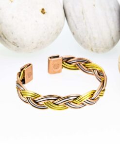 Braided magnetic copper therapy bangle combining copper, brass, and silver-tone strands, with rectangular magnetic ends, displayed against a backdrop of smooth white pebbles.