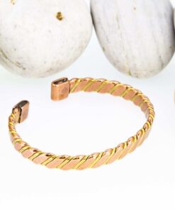 Twisted two-tone magnetic copper therapy bangle with a copper and brass spiral design, finished with rectangular magnetic ends, displayed against smooth white pebbles.