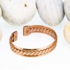 Magnetic copper therapy bracelet in an open cuff style, featuring a polished copper band with a central braided pattern, smooth edges, and magnets embedded at each end for potential arthritis relief, improved circulation, and overall wellness, displayed against smooth white pebbles.