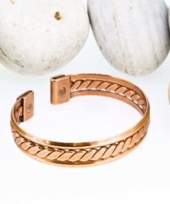 Magnetic copper therapy bracelet in an open cuff style, featuring a polished copper band with a central braided pattern, smooth edges, and magnets embedded at each end for potential arthritis relief, improved circulation, and overall wellness, displayed against smooth white pebbles.