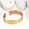 Magnetic copper therapy bracelet in an open cuff design, featuring a smooth polished copper base accented with parallel brass stripe inlays, with magnets embedded at each end for potential arthritis relief, improved circulation, and holistic wellness benefits, displayed against smooth white pebbles.