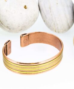 Magnetic copper therapy bracelet in an open cuff design, featuring a smooth polished copper base accented with parallel brass stripe inlays, with magnets embedded at each end for potential arthritis relief, improved circulation, and holistic wellness benefits, displayed against smooth white pebbles.