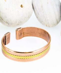 Magnetic copper therapy bracelet in an open cuff style, featuring a polished copper band with a single central brass inlay in a fine zigzag pattern, complemented by magnets at each end for potential arthritis relief, enhanced circulation, and overall wellness, displayed against smooth white pebbles.