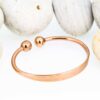 Rose gold-coloured open copper therapy bangle with a smooth polished finish, featuring two magnetic ball ends, displayed against a backdrop of smooth white pebbles.