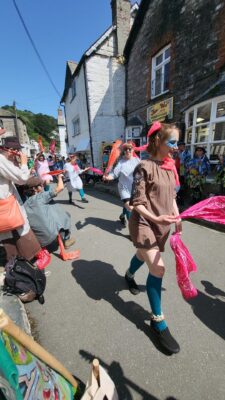 The Wad morris dancers performing on a sunny day in a narrow coastal village street, right outside the historic Joan the Wad Shop in Polperro. The dancers are wearing bright ribbons, teal socks, and waving pink and orange bandanas.