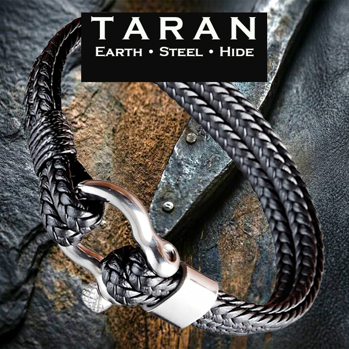 A Taran men's bracelet with a black braided leather band and a polished silver steel shackle clasp, displayed over rugged grey slate and old wood. The logo text 'TARAN Earth Steel Hide' is in the top right.