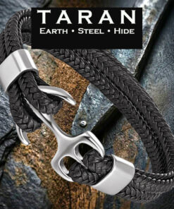 A Taran men's black braided leather anchor bracelet featuring a wide multi-strand layout. Multiple thick rows of tightly woven black leather hide run parallel to each other, forming a dense, tactile herringbone texture. The strands wrap through a large, stylized, mirror-polished silver stainless steel maritime anchor that functions as a hook clasp, secured by wide silver steel retention collars. The bracelet is shot at a sharp angle over a dark, texturized slate rock and a piece of weathered wood, beneath the black Taran brand logo with the tagline Earth Steel Hide.