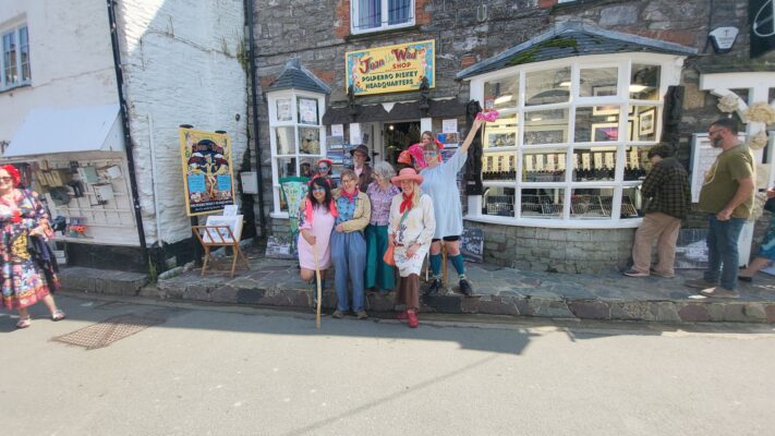 A lively group of traditional street performers posing for a photo directly in front of the entrance to the Joan the Wad Shop in Polperro on a sunny day.