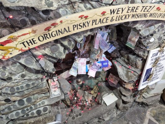 Stone grotto wishing well inside The Original Pisky Place shop in Polperro, embedded with coins, hanging banknotes, red fairy lights, and Joan the Wad signs.